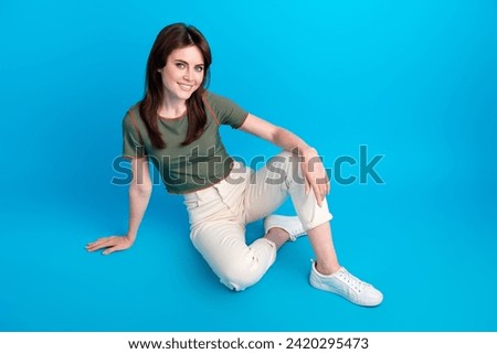 Full length photo of optimistic pleasant adorable girl dressed khaki t-shirt sit on floor smiling isolated on blue color background