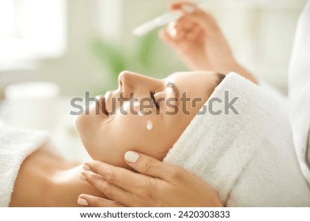 Close up beautician's hands applying anti-aging facial cream on woman client face to prevent wrinkles in spa salon. Skin care, cosmetic procedures for facial care and beauty treatment concept. Royalty-Free Stock Photo #2420303833