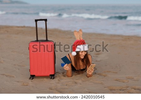 Happy young woman in Christmas Santa Claus hat lying on sand seashore with red suitcase holds smartphone, credit card and passport looks at camera. New Year winter holiday vacation at tropical resort.
