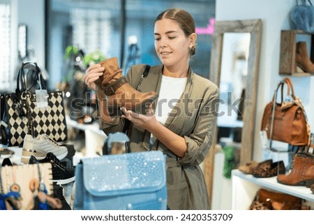 Girl shopper in leather product store finds it difficult to choose and thoughtfully view leather shoe boots near showcase with winter shoes. Smiling attractive young woman shopping at shoes store