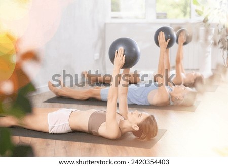 Motivated young woman maintaining active lifestyle exercising with ball during group workout in modern fitness center..