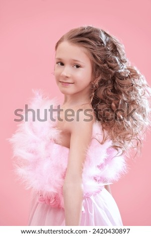 Children's holiday fashion. Beautiful little lady girl with lush curly hair and in a pink evening dress with feathers smiles. Pink studio background. Little Princess.