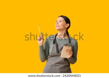 Beautiful young waitress with notebook and pencil on yellow background