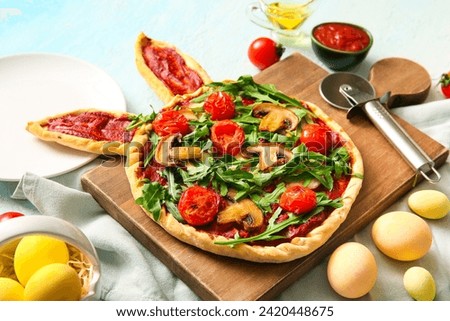 Wooden board with tasty pizza and painted eggs for Easter celebration on blue background