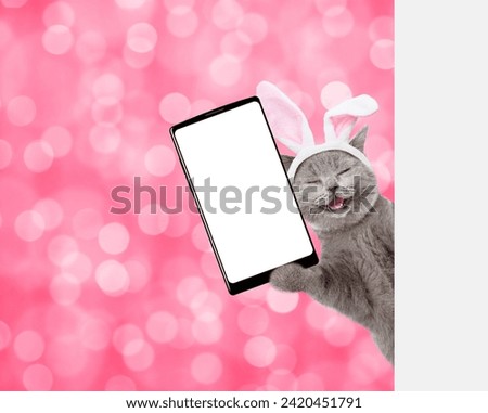 Happy kitten wearing easter rabbits ears holds big smartphone with white blank screen and looks from behind empty white banner on pink blurred background