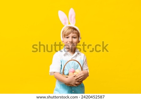 Cute little boy in bunny ears with basket of Easter eggs on yellow background