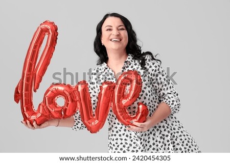 Beautiful happy mature woman with balloon in shape of word LOVE on grey background. Valentine's Day celebration