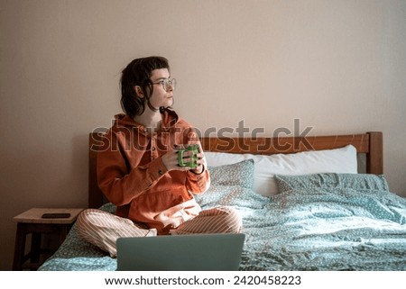 Woman freelancer drinking tea having break sitting in bed at home looking at window. Female in glasses pyjama working. Freelance, remote, distant work job in relaxed atmosphere, self-organization.  Royalty-Free Stock Photo #2420458223