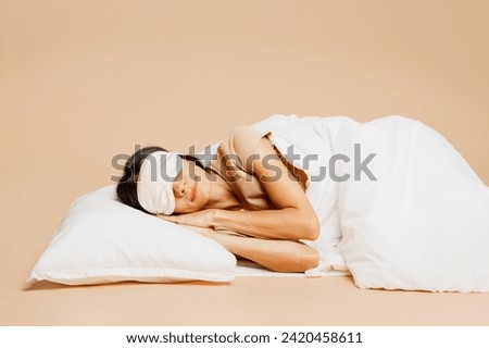 Asleep side view young calm Latin woman she wear pyjamas jam sleep eye mask rest relax at home lay down under duvet close eyes isolated on plain pastel beige background. Good mood night nap concept