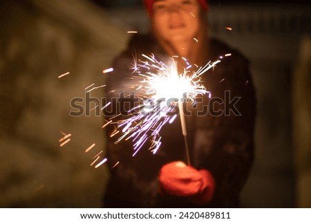 Woman holding sparkler night while celebrating Christmas outside. Dressed in a fur coat and a red headband. Blurred christmas decorations in the background. Selective focus
