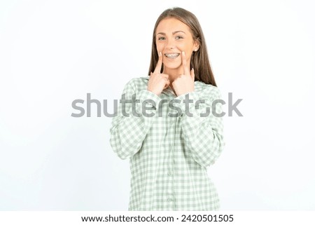Happy Young beautiful woman wearing green plaid pyjama with toothy smile, keeps index fingers near mouth, fingers pointing and forcing cheerful smile