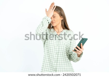 Upset depressed Young beautiful woman wearing green plaid pyjama makes face palm as forgot about something important holds mobile phone expresses sorrow and regret blames