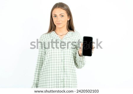 Young beautiful woman wearing green plaid pyjama holds new mobile phone and looks mysterious aside shows blank display of modern cellular