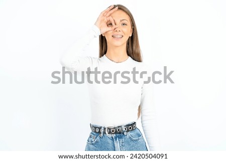 Young beautiful woman standing over white studio background doing ok gesture with hand smiling, eye looking through fingers with happy face.