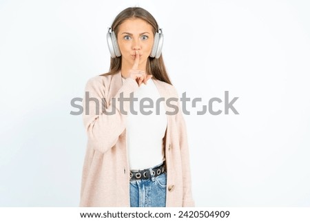 Young beautiful woman standing over white studio background making hush gesture with finger on her lips wearing  wireless headphones. Be quiet. Royalty-Free Stock Photo #2420504909