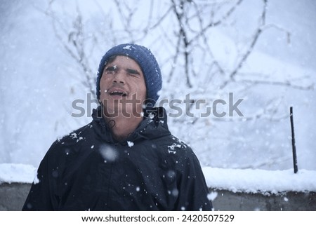 Picture of Cheerful man enjoying snowflakes falling from upwards, picture of adult indian boy with snow backround
