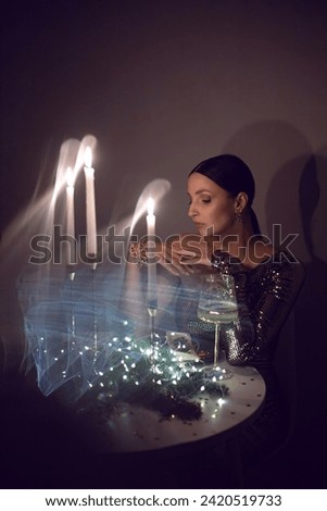 Portrait of a girl in a gold festive sequined dress sitting at a round table at home on Christmas night. On the table there are glasses with wine and branches of fir tree with garland