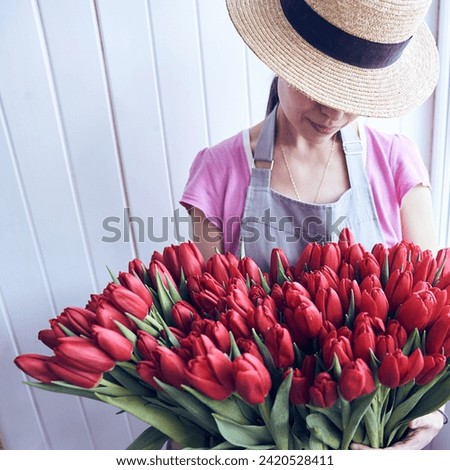 Square image of unrecognisable woman holding a big bouquet of red tulips with a copy space. Gardener wearing a hat, apron and holding beautiful flowers. 