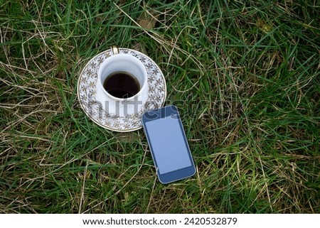 A mobile phone and a cup of coffee on a saucer with gold patterns are standing on the grass in a clearing. Image about remote work in nature, for your creative design or illustrations.