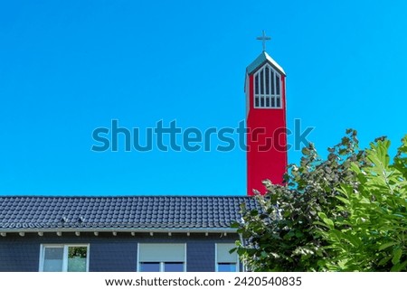Roof of a Christian church with a red church tower and a cross over green trees against a blue sky with clouds.