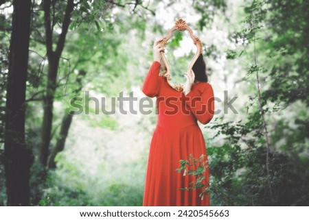 woman in red dress covers her face with a mirror that reflects the surrounding forest Royalty-Free Stock Photo #2420545663