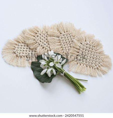 Four beige heart-shaped macramé coasters form a semi-circle on a white table, highlighting a central bouquet of white snowdrops. A simple yet elegant composition. Royalty-Free Stock Photo #2420547551
