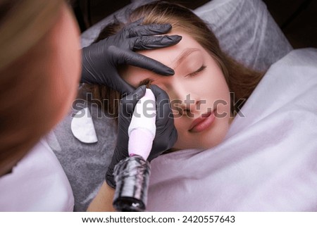 In the hands of the master tattoo machine he applies permanent makeup on the eyebrows. PMU Procedure, Permanent Eyebrow Makeup. Royalty-Free Stock Photo #2420557643