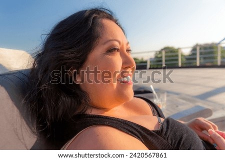 Profile portrait of Mexican Latina woman sitting on sun lounger at stern of cruise ship, blurred background, beautiful smile, long black hair, light makeup and low-cut dress, enjoying a sunny day