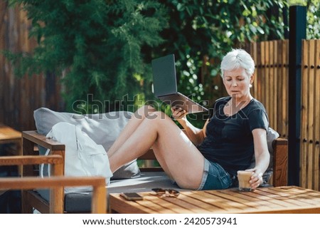 Adult woman with blonde hair using laptop on back yard garden. Working from home.