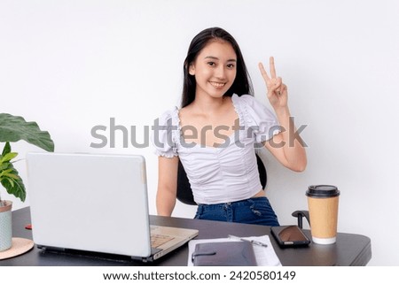 A smiling young female college student shows a peace sign while studying at her desk with a laptop, coffee, and smartphone.