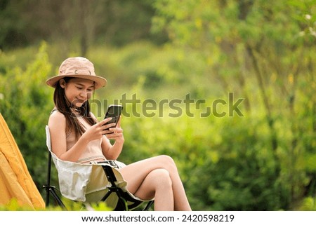 young asian woman sitting in a chair nex to tent using her phone on hand at campground,
