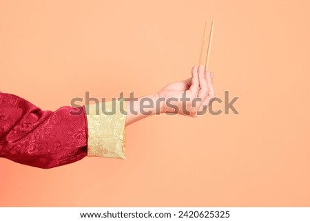 Happy Chinese new year. Hand of asian chinese senior man wearing red traditional cheongsam qipao or changshan dress with gesture of hand holding wood chopsticks isolated on orange background.