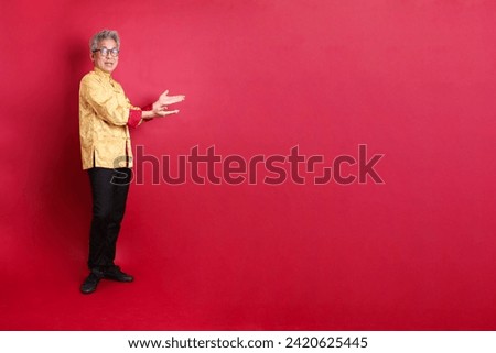 Happy Chinese new year. Asian Chinese energetic senior man wearing golden traditional cheongsam qipao or changshan dress with gesture of pointing isolated on red background.