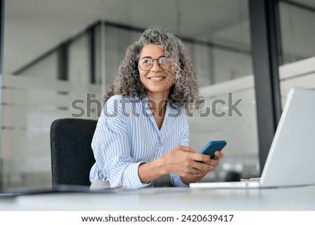Happy mature business woman executive holding mobile cell phone looking away in office. Smiling middle aged professional businesswoman manager wearing glasses using cellphone working on smartphone. Royalty-Free Stock Photo #2420639417