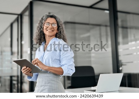 Smiling mature professional business woman bank manager, older happy female executive or lady entrepreneur holding digital tablet pad standing in office at work, looking away at copy space. Royalty-Free Stock Photo #2420639439