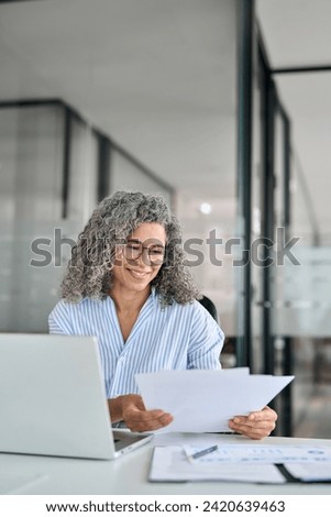 Busy middle aged business woman working in office with laptop reading document. Mature professional hr manager, notary holding paper finance report sitting at desk in office. Consultancy. Vertical. Royalty-Free Stock Photo #2420639463