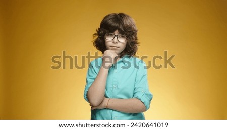Cute caucasian boy in glasses looking at camera and thinking about something, isolated on yellow background - emotions concept close up 