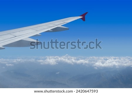 View of the wing from a window in the passenger cabin of an airplane. There are mountains on the ground and snow-capped mountains in the distance.