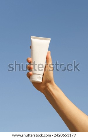 Vertical image of a female hand holding a white mockup tube of cream on a blue sky background. Concept of natural cosmetics for face and body, beauty products Royalty-Free Stock Photo #2420653179