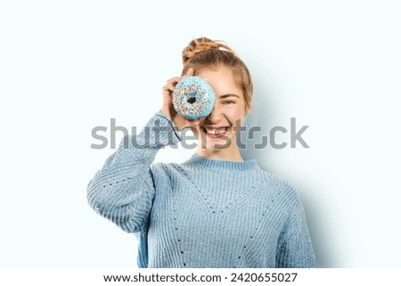 Young positive smiling laughing woman covers one eye with a blue donut, having fun, playing with sugary doughnut on a blue background, sweets, junk food, fast food concept, cheat meal day