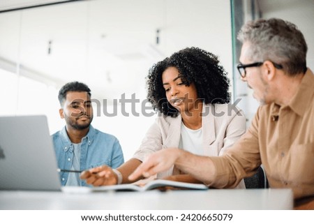 Focused businesswoman in chic blazer leans over to discuss a document with her colleagues, demonstrating teamwork and active problem-solving in a contemporary office. Work-team solving tasks together Royalty-Free Stock Photo #2420665079