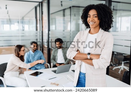 Smart high-skilled young Brazilian woman with a cheerful smile stands with arms crossed on the forefront in a modern office setting with her colleagues collaborating in the background, team synergy Royalty-Free Stock Photo #2420665089