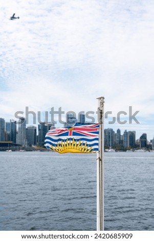 A British Columbia Flag flapping in the wind with the Vancouver Skyline and a Seaplane in the background.
