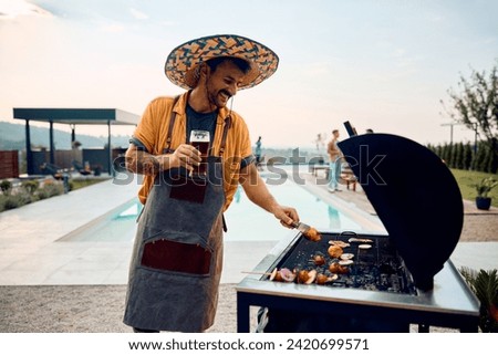 Happy man preparing barbecue while having a glass of beer during poolside party with friends.