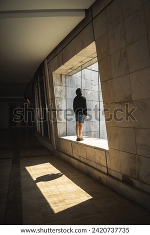 Young man in black hoodie and blue shorts stands in shadowed corridor at Cidade da Cultura in Santiago de Compostela. Sunlight streams through square opening, casting captivating shadow play. Royalty-Free Stock Photo #2420737735