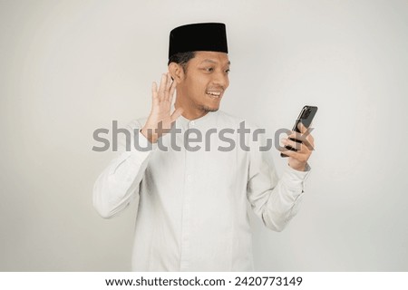 Happy smiling Asian Muslim man standing with Eid Mubarak greeting gesture and welcoming Ramadan video call via mobile phone