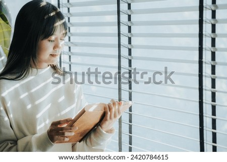 A young Asian woman is sitting by the window in a cafe reading a book.