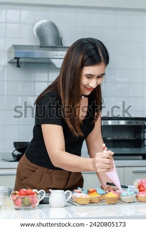 Asian professional female pastry bakery baker chef housewife standing using tube bag piping squeezing delicious pink whipped cream decorating topping preparing cupcakes in full decorated kitchen.