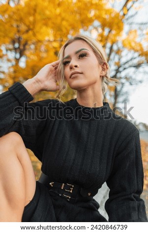 Fashionable beautiful fresh woman with clear skin face in a fashion black knitted dress sits in an autumn park with yellow foliage outdoors