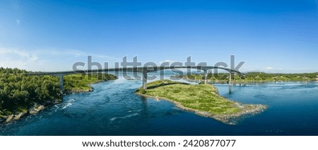 A panoramic drone shot of Saltstraumen Bridge arching over the world's strongest tidal current, with lush green islets and distant mountains under a clear blue sky